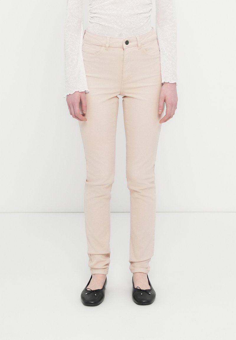 Γυναίκα που φοράει εφαρμοστά ανοιχτό μπεζ παντελόνια, λευκή μπλούζα μακρυμάνικη από δαντέλα και μαύρα μπαλαρίνες, στέκεται μπροστά σε απλό λευκό φόντο.