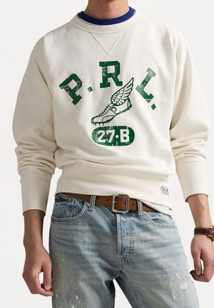 Homme portant un sweat crème avec texte vert "P.R.L.", graphique de sneaker ailée et numéro "27·B", associé à un jean bleu et une ceinture marron.