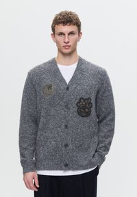 Grå strikket cardigan med v-hals, frontknapper og to lapper: en med et globusdesign og en anden med et stiliseret ansigt. Blød tekstur.