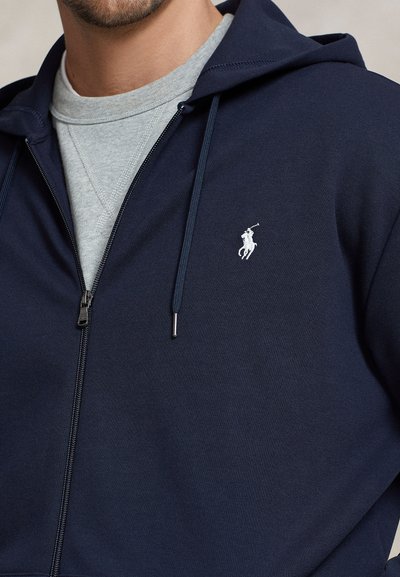 Tmavě modrá mikina na zip z měkké látky s kapsou kangaroo, nastavitelnou kapucí se šňůrkami a malým bílým logem polovina na hrudi.