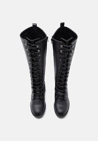 Bottes noires en similicuir montant jusqu'au genou avec un laçage complexe, des œillets argentés et des languettes en haut pour un enfilage facile. Texture lisse sur toute la surface.