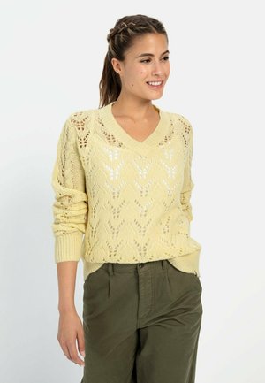 Gelber, gestrickter Pullover mit V-Ausschnitt und offenen Rautenmuster. Lange Ärmel mit gerippten Bündchen, kombiniert mit olivgrünen Hosen.