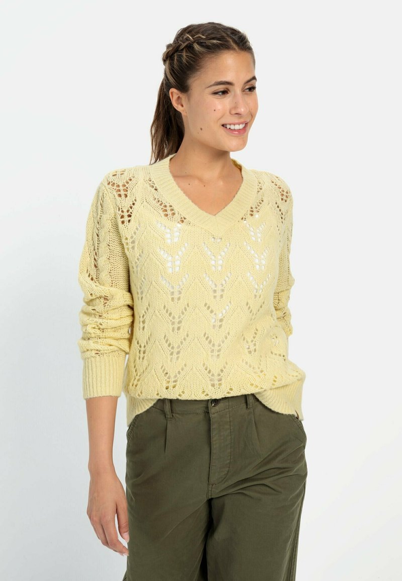 Gelber, gestrickter Pullover mit V-Ausschnitt und offenen Rautenmuster. Lange Ärmel mit gerippten Bündchen, kombiniert mit olivgrünen Hosen.