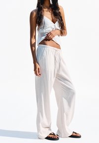 Vitt linnekroppat linne med snurrdetalj och lösa, ljusbeige byxor. Svarta flip-flops fulländar outfiten. Klar vit bakgrund.