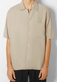 Short-sleeve button-up shirt in a light beige fabric. Notable features include a chest pocket, soft texture, and a classic collar design.