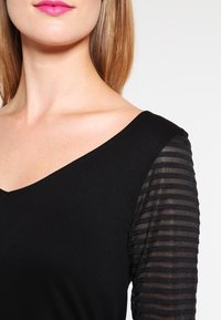 Schwarzes Oberteil mit V-Ausschnitt, unifarbenem Oberteil und transparenten gestreiften langen Ärmeln. Glatte Textur mit figurbetontem Design.