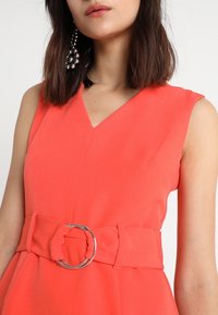 Korallfärgad ärmlös klänning med djup V-ringning och bälte i midjan som har en silverfärgad rund spänne. Slät tygkänsla, inga mönster.