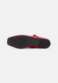 Zapato de charol rojo con una superficie lisa, punta afilada y suela de goma con un patrón estriado para mayor agarre. Acabado minimalista en dorado.