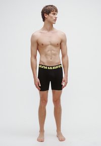Bóxers negros con una cinturilla ancha de color amarillo brillante que presenta el logo "DSQUARED2". El modelo está sin camiseta y de pie, descalzo.
