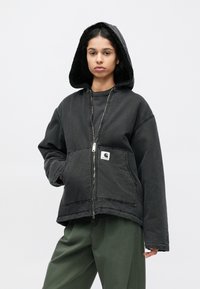 Melna kapuce ar rāvējslēdzēja aizdari, ar teksturētu oderi, priekšējām kabatām un logotipa plāksteri. Modelis valkā zaļas vaļīgas bikses.