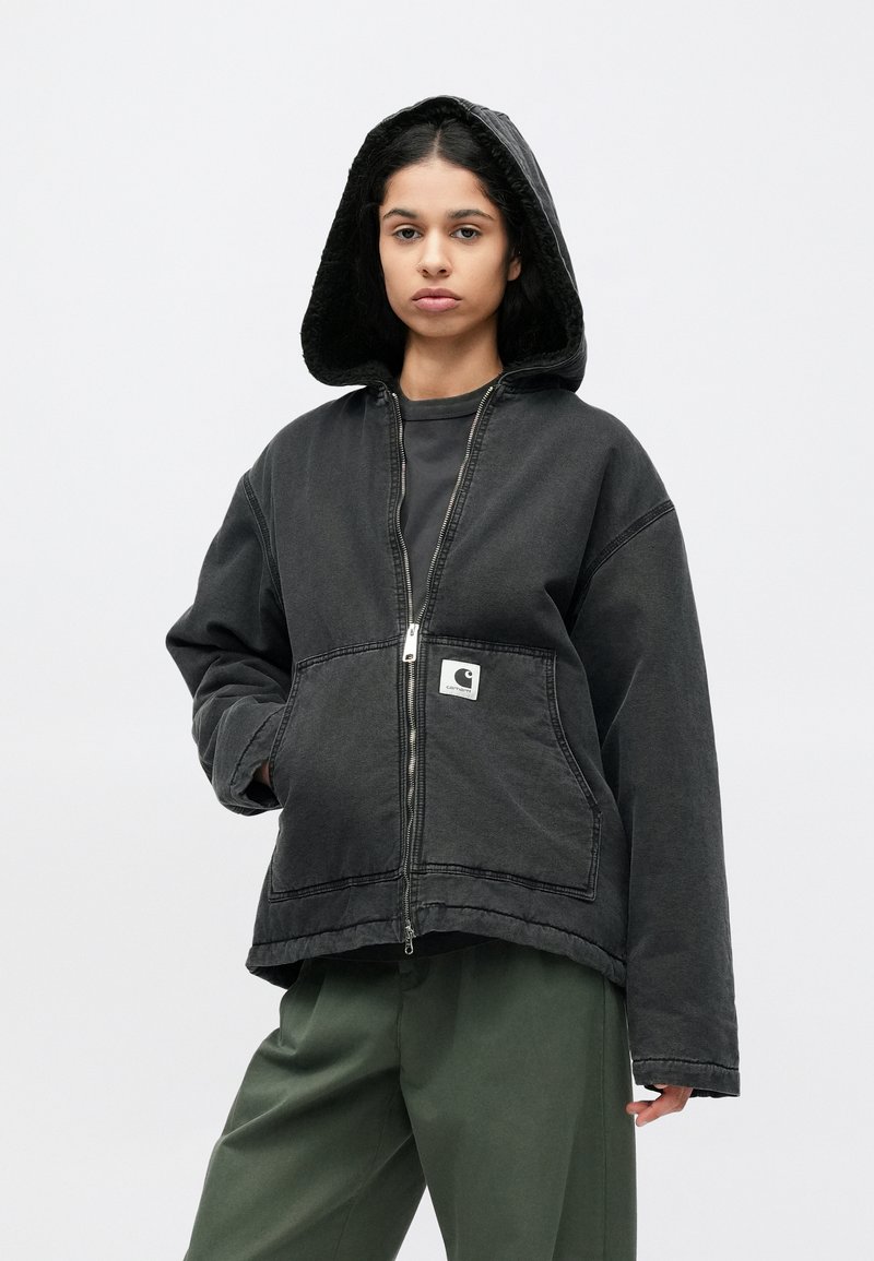 Zwarte capuchontrui met ritssluiting, voorzien van een gestructureerde voering, voorzakken en een logopatch. Model draagt losse groene broek.