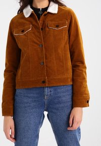 Bruine corduroy jas met witte schapenvachtkraag, voorzien van twee borstzakken, knoopsluiting en een getailleerde pasvorm. Gecombineerd met blauwe spijkerbroek.