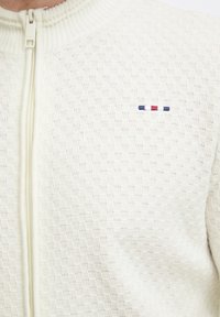 Cremefarvet lynlånsweater med et struktureret mønster, der har et lille broderet logo i rød, hvid og blå nær kraven.