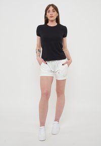 Maglietta nera a maniche corte abbinata a pantaloni corti bianchi elasticizzati con tasche frontali e un logo, indossati con sneakers bianche.