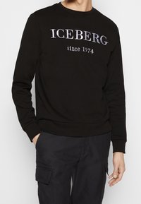 Felpa nera con scollatura a girocollo, caratterizzata dal testo "ICEBERG dal 1974" in argento. Tessuto morbido con maniche lunghe e polsini a costine.