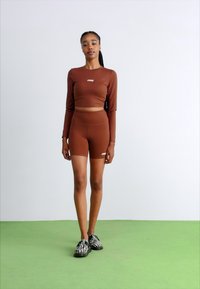 Brun gli langermet topp med matching høy midje shorts. Modellen har på seg svart og hvit zebra-stripede sko. Enkelt bakgrunn.