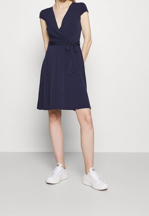 Kobieta ubrana w granatową sukienkę o długości do kolan z krótkimi rękawami i białe sneakersy na platformie, stojąca na tle gładkiego jasnego tła.