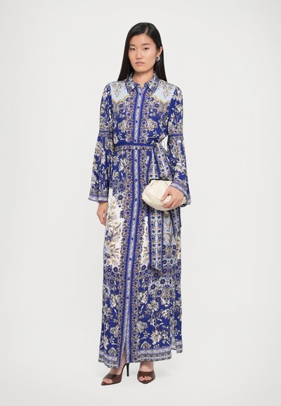 Robe longue à motifs floraux bleus avec des accents blancs et dorés, colée, cintrée à la taille, manches longues en forme de cloche, associée à un sac crème.