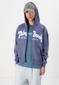 Lilla zip-up hættetrøje med hvid "Palm Beach Florida" grafik, lagd over en blå skjorte, parret med lysegrå bukser og en grå kasket.