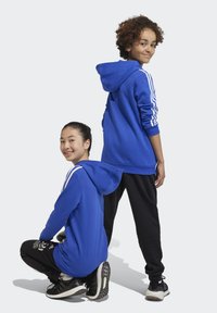 Dos niños que visten sudaderas azules a juego con rayas blancas en las mangas. Uno está arrodillado, mientras que el otro está de pie con pantalones jogger y zapatillas negras.