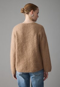 Kvinne med hår i knute, iført sølvøredobber, en puffy lysebrun genser og blå jeans, vendt bort fra kameraet mot en enkel grå bakgrunn.