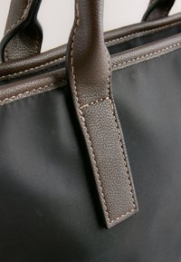 Gros plan d'un sac en tissu noir avec des poignées en cuir marron foncé et des coutures détaillées sur les anses et les bords.
