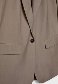 Blazer beige avec une fermeture à un seul bouton noir, deux poches à rabat à l'avant et un col à revers ajusté.
