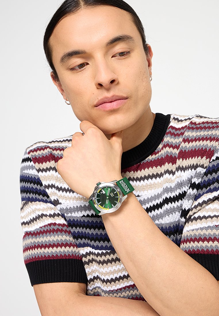 Reloj plateado con una esfera verde y negra, correa de silicona verde, con un acento amarillo. Suéter de patrón multicolor con ribete negro.