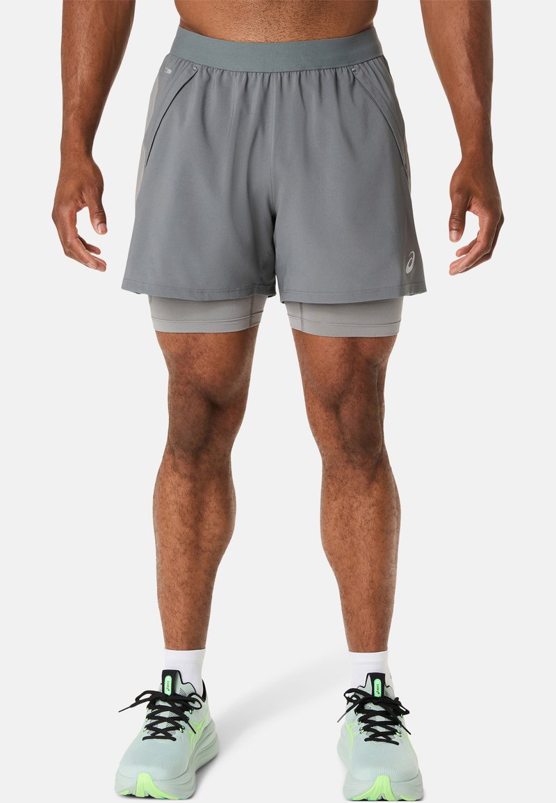 Shorts de sport gris avec une ceinture et des poches latérales, portés avec des shorts de compression gris clair en dessous. Chaussures de sport vert fluo.