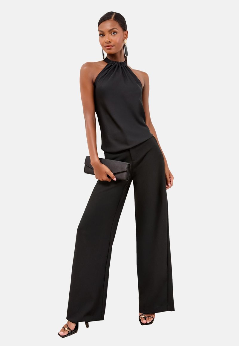 Femme portant un top noir sans manches à col halter, un pantalon noir à jambes larges, des sandales noires à talons hauts, tenant une pochette noire.