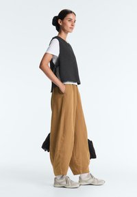 Femme de profil portant un gilet noir sans manches, une chemise blanche, un pantalon ample beige, des chaussettes blanches et des baskets beiges, les mains dans les poches.