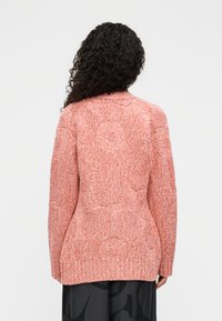 Γυναίκα με σγουρά μαλλιά που φοράει πλεκτό πουλόβερ με μακριά μανίκια σε πορτοκαλί χρώμα με υφή και φαρδιά μαύρη παντελόνα με σχέδια, φαντασμένη από πίσω.