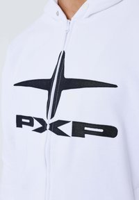 Sudadera blanca con cremallera y capucha que presenta un gran logotipo negro bordado con líneas definidas y el texto "PXKP". Tejido suave con un diseño ajustado.