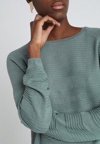 Pull vert texturé avec nervures horizontales et encolure ronde. La main du mannequin repose sur le menton, mettant en valeur un design simple.