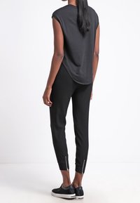 Person som bär en svart ärmlös topp och smala svarta byxor med anklarade dragkedjor, står vänd bort från kameran mot en enfärgad vit bakgrund.