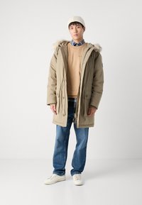 Beige parka takki, jossa on karvavuorattu huppu, päällä beige collegepaita ja ruudullinen paita. Siniset farkut ja valkoiset lenkkarit täydentävät asun.
