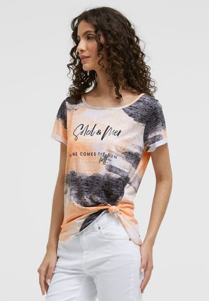 Woman with curly hair wearing a multicolored short-sleeve T-shirt with text and white pants, standing against a plain background.