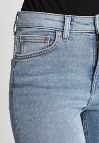 Acercamiento de unos pantalones de mezclilla azul claro en la cintura y la parte superior del muslo, con un bolsillo frontal con remaches de cobre y trabillas para el cinturón, combinados con una blusa negra.