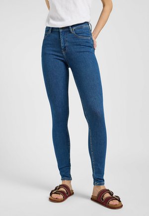Jeans ajustados de mezclilla de color azul, con una cintura de altura media, diseño de cinco bolsillos y detalles de costura sutiles. Se combinan con sandalias marrones de tipo deslizante.