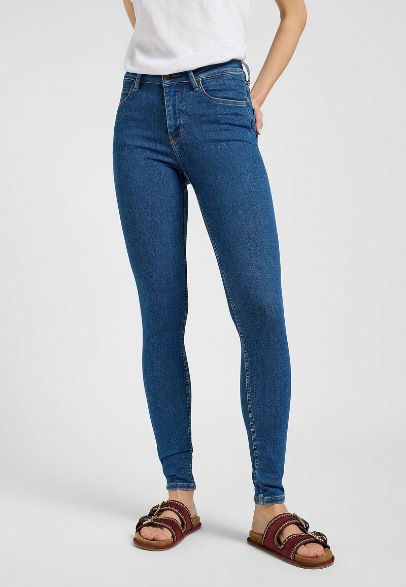 Jeans ajustados de mezclilla de color azul, con una cintura de altura media, diseño de cinco bolsillos y detalles de costura sutiles. Se combinan con sandalias marrones de tipo deslizante.