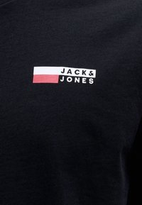 Μαύρο ύφασμα με μικρό λογότυπο που δείχνει "JACK & JONES" σε λευκά γράμματα με λευκά και κόκκινα ορθογώνια φόντου.