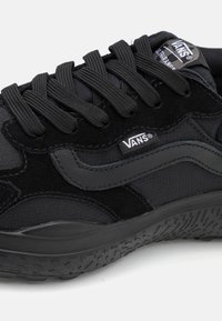 Des baskets Vans noires en suède et tissu texturé, avec une semelle en caoutchouc épaisse et un design empilé. Logo emblématique sur le côté.