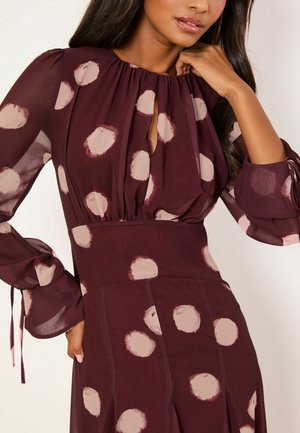 Naine kannab tumevürtspunast kleiti, millel on suured ebaühtlased heleroosad täpid, pikad läbipaistvad varrukad ja võtmekohaga kaelusdetail.