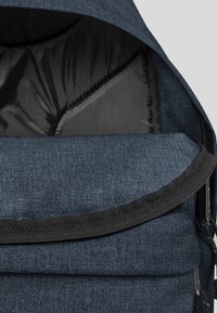 Mochila de tela azul oscura con cierre de solapa, exterior texturizado, interior acolchado y acentos negros. Cuenta con una cremallera y tirantes ajustables.