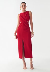 Red midi dress with a fitted bodice and twist detail at the waist. Features a side slit and smooth texture. Paired with strappy heels.