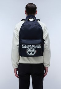 Zaino navy con una grande tasca anteriore. Presenta un logo e testo bianchi, spallacci regolabili e un tessuto con una superficie liscia.