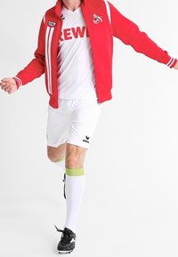 Veste à fermeture éclair rouge avec des rayures blanches et un logo, maillot blanc avec le texte « CREW », shorts blancs, chaussettes accentuées de vert, et chaussures de football noires.