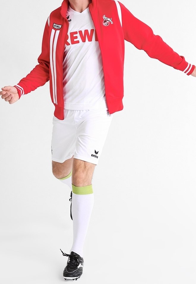 Veste à fermeture éclair rouge avec des rayures blanches et un logo, maillot blanc avec le texte « CREW », shorts blancs, chaussettes accentuées de vert, et chaussures de football noires.