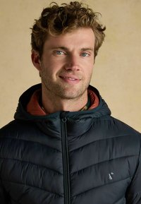 Młody mężczyzna z kręconymi włosami ma na sobie czarną kurtkę puchową z kapturem i czerwoną podszewką, uśmiecha się na tle jednolitego beżowego tła.