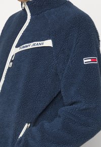 Námořnická modrá fleecová bunda s vysokým límcem, bílým zipem a bílým logem "TOMMY JEANS". Na rukávu se nachází malé vícerozměrné logo.